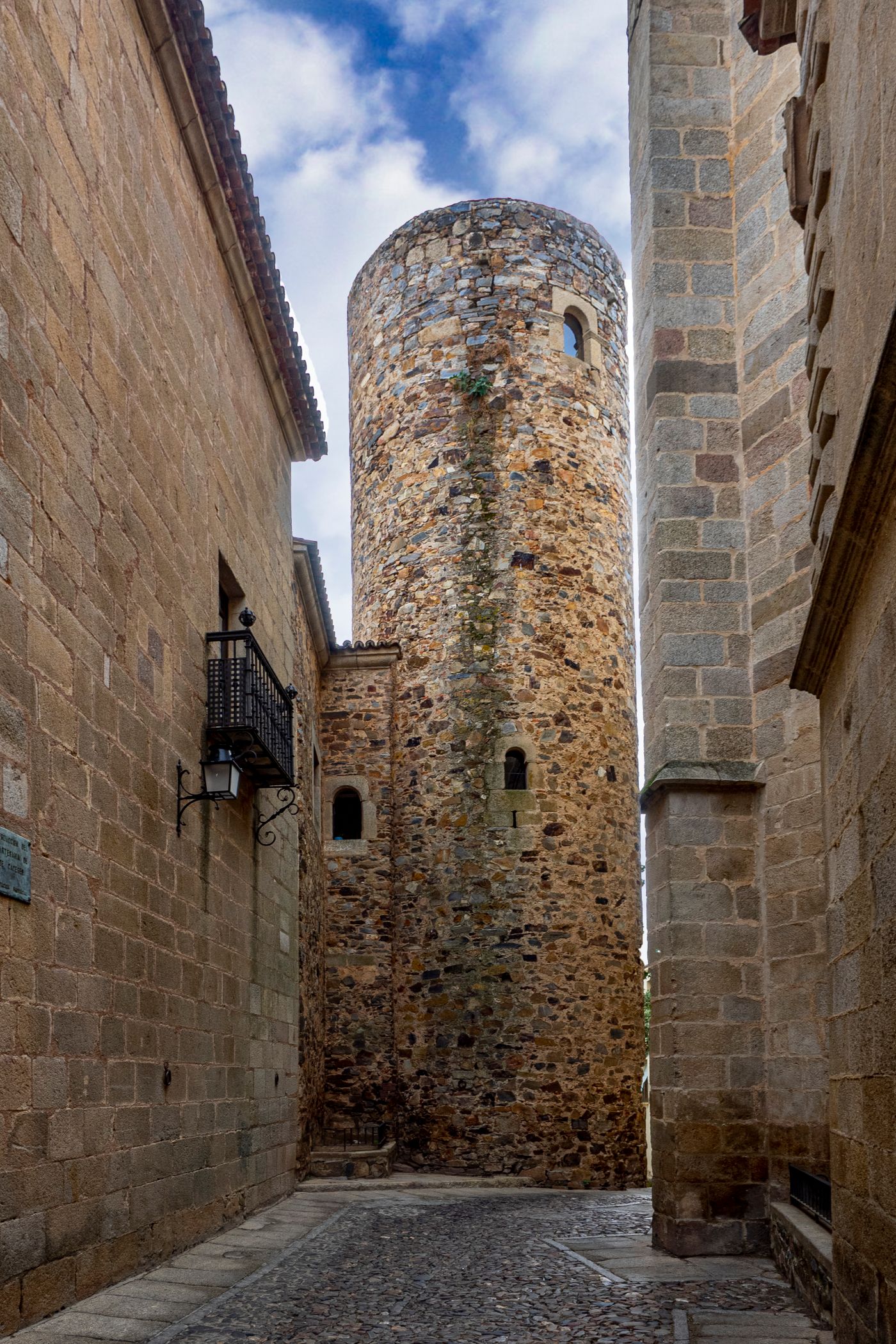 Calle empedrada entre muros de piedra con una torre al fondo
